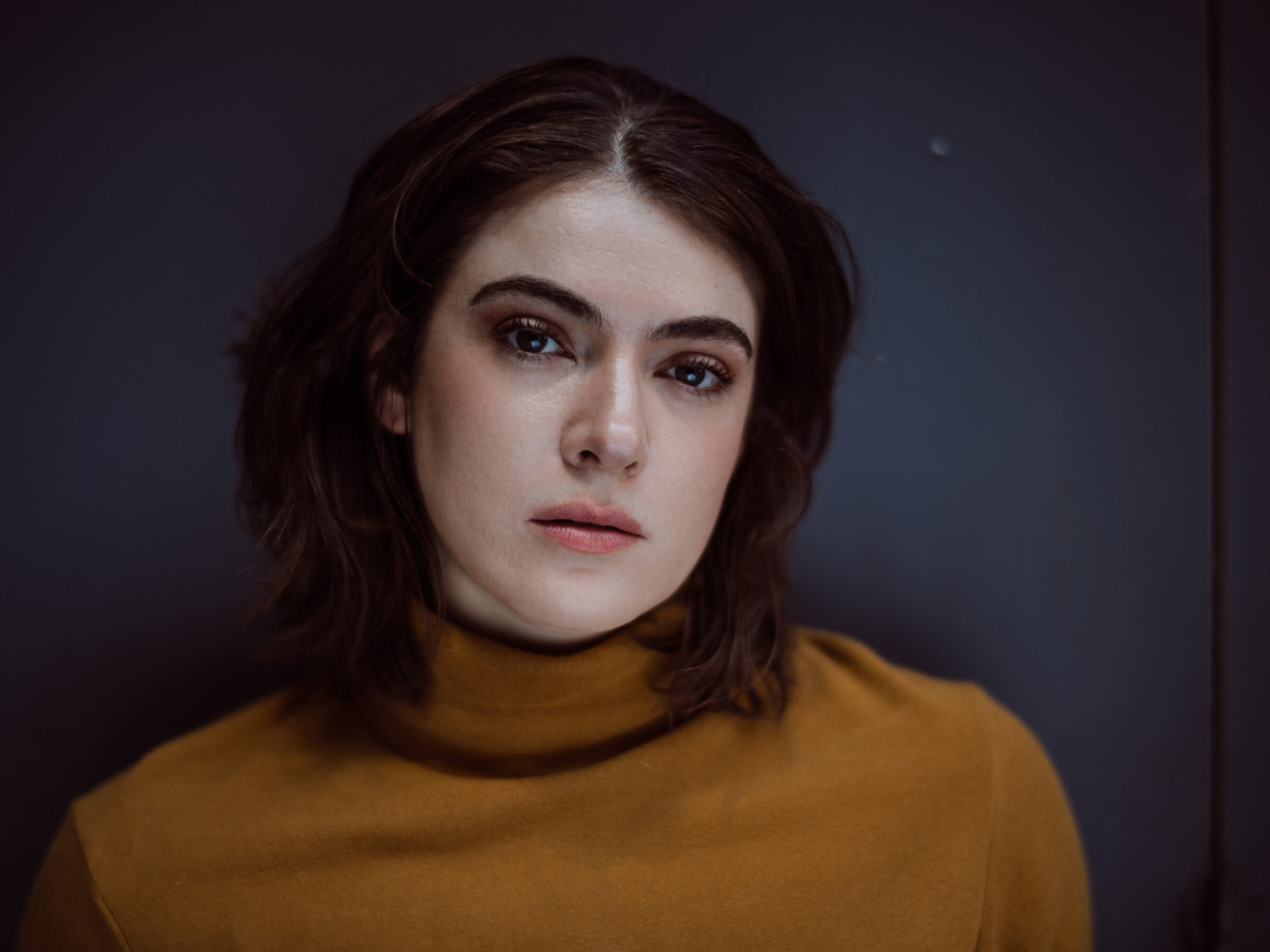 A photo of Meg in a mustard yellow skivvy, looking into the camera.